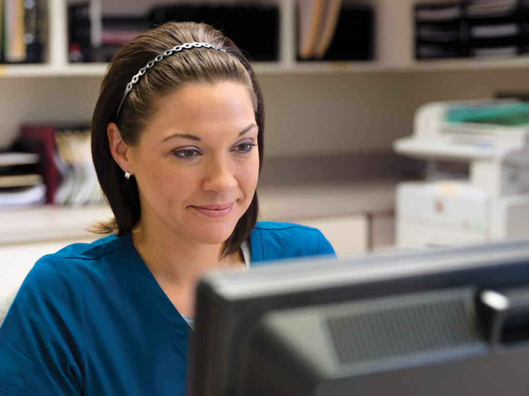 A nurse in blue scrubs smiles while working at a computer workstation in a medical office setting.