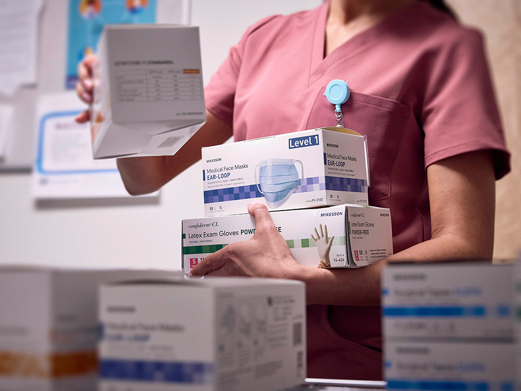A healthcare worker in pink scrubs carries stacked boxes of McKesson medical supplies, including face masks and latex exam gloves.